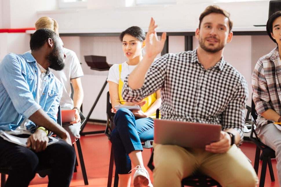 a group of people sitting on the chairs in a room, one raises the hand