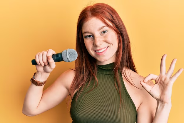 Young red head girl singing song using microphone doing ok sign with fingers